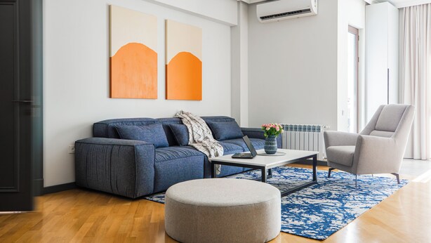 Ein modernes Wohnzimmer mit einer Couch, einem Hocker und einem Sessel, einem Couchtisch mit einem Laptop sowie zwei abstrakten Gemälden an der Wand.