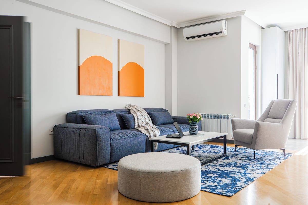 Un salon moderne avec un canapé, un pouf et un fauteuil, une table basse avec un ordinateur portable, et deux tableaux abstraits accrochés au mur.