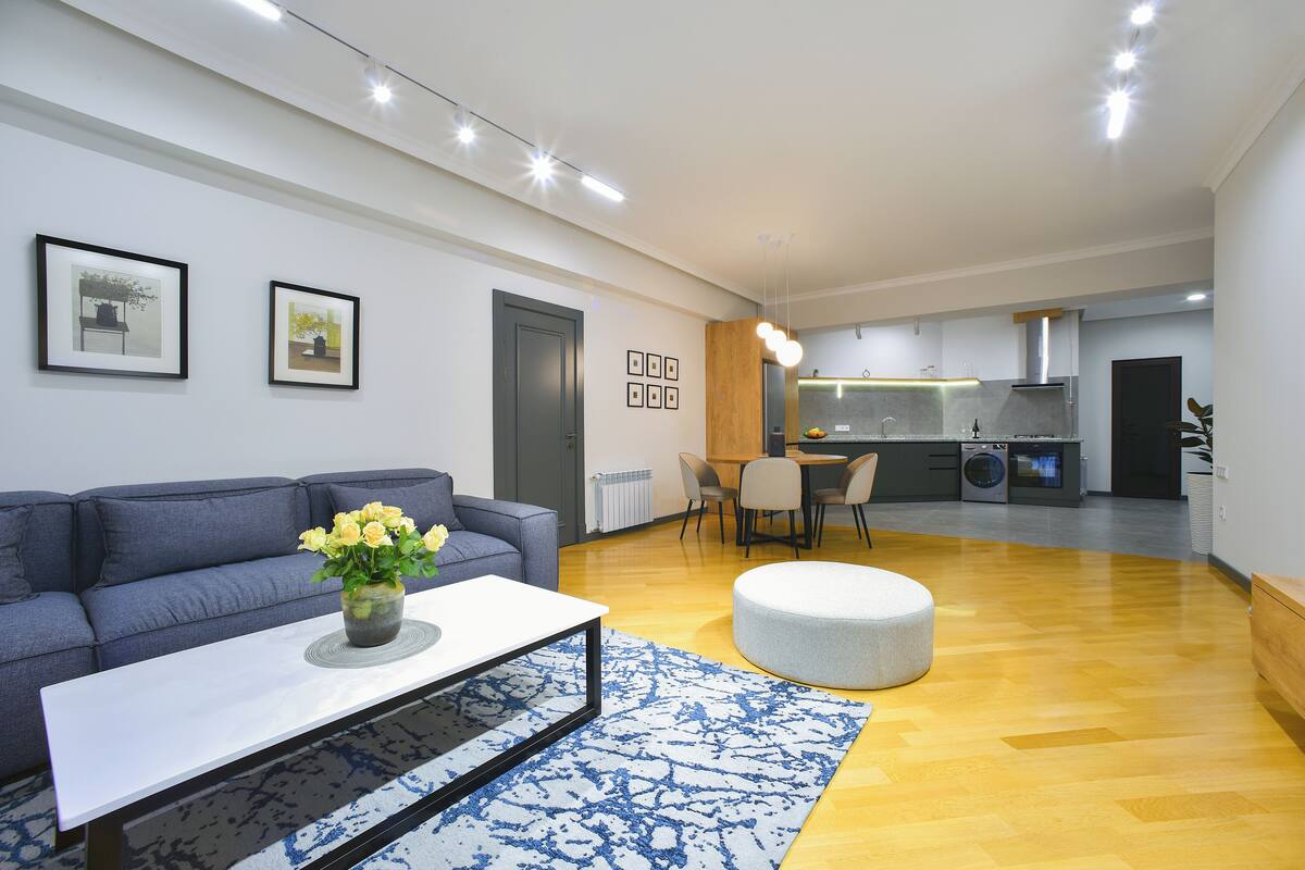 Un salon moderne avec un canapé, une table basse et un pouf. On aperçoit, au fond, un coin cuisine avec une table à manger et des chaises.