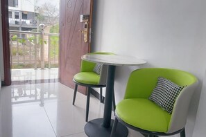 Een kamer met een groene stoel en een klein tafeltje, een houten deur en uitzicht op een balkon met een hekwerk.