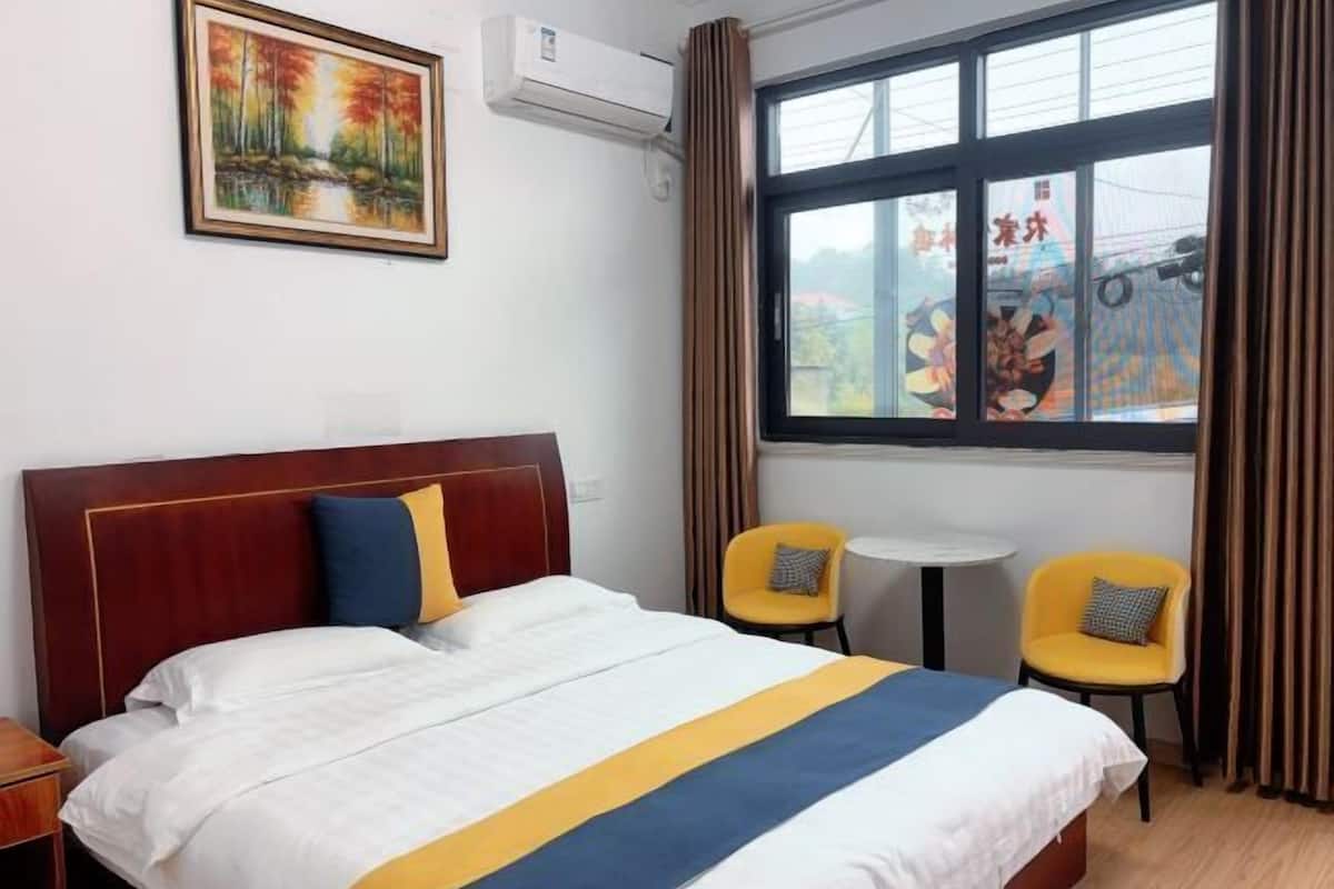 Een hotelkamer met een bed, een schilderij, een airconditioner, een raam met gordijnen en twee stoelen.
