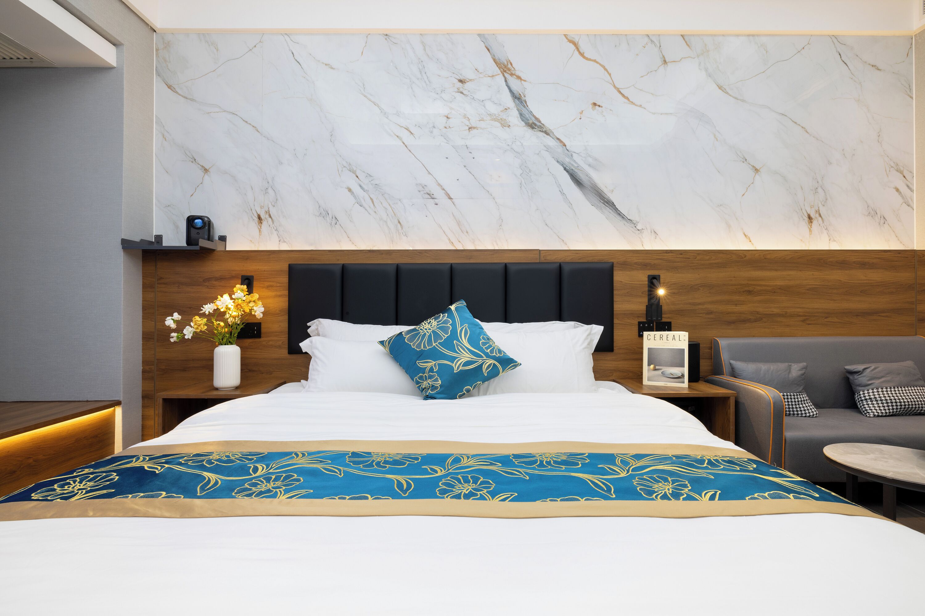 Una habitación de hotel con pared de diseño marmolado, una cama con colcha floral azul, cabecera de madera y sofá gris.