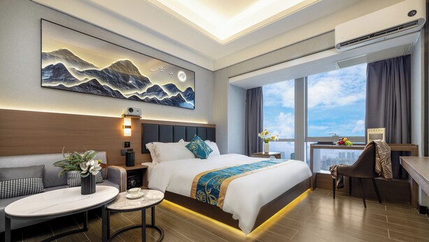 Una habitación de hotel moderna con una cama grande, un escritorio, una mesa redonda y vistas a la ciudad a través de amplias ventanas.
