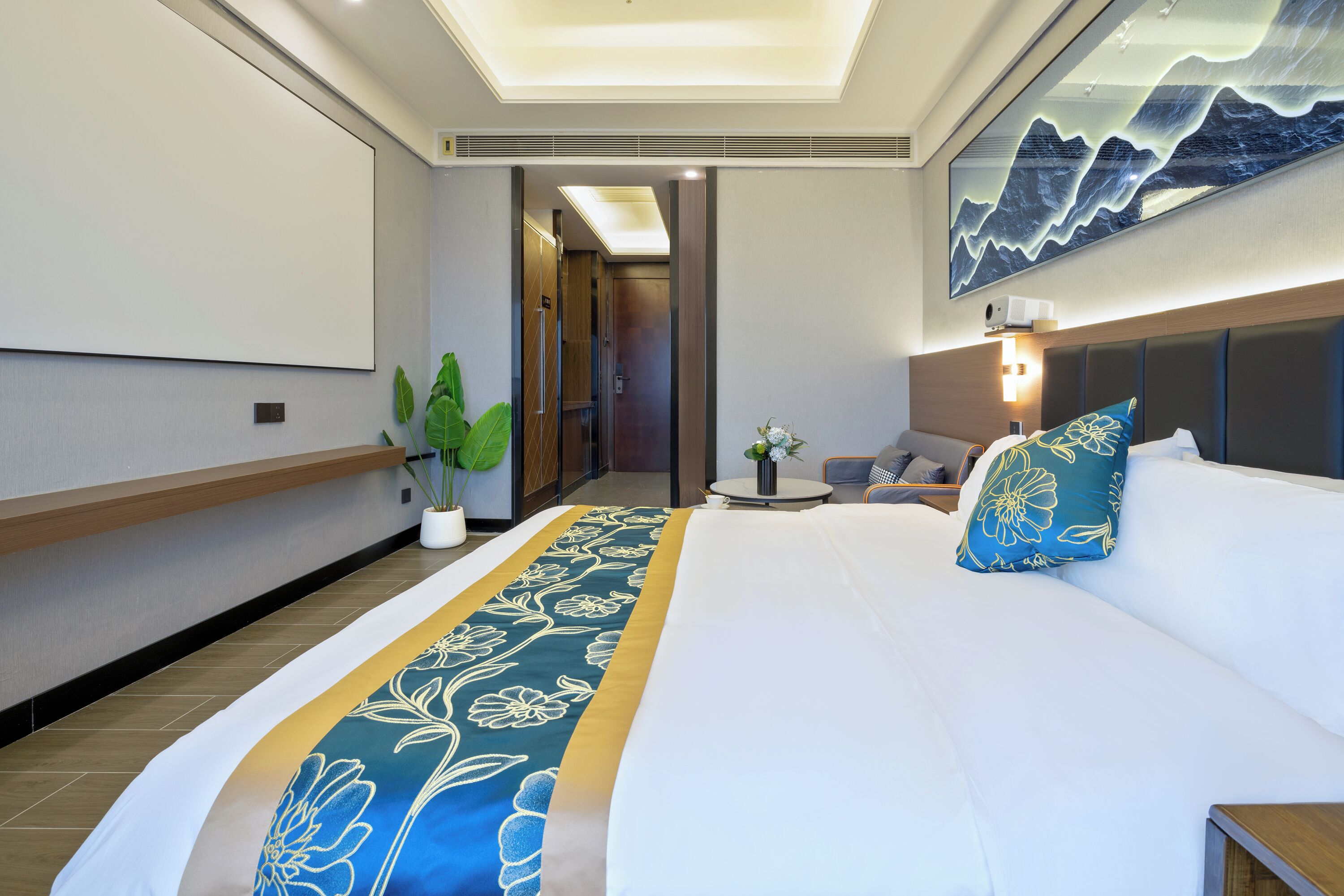 Une chambre d’hôtel avec un grand lit, une table de chevet, une plante en pot et un téléviseur fixé au mur.