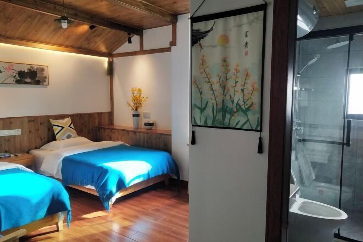 A room with two beds, wooden flooring, a painting on the wall, and a glass shower enclosure.