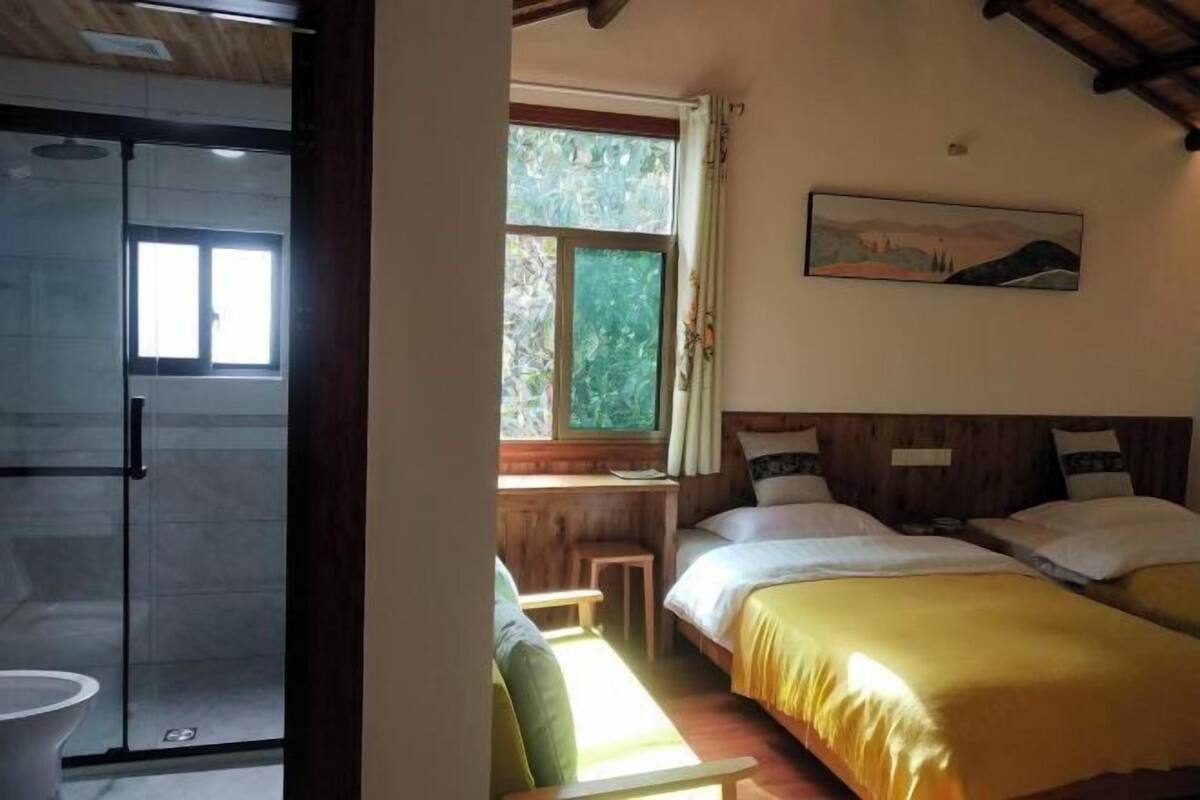 A room with two beds, a wooden ceiling, a window with a view of greenery, and a painting on the wall.