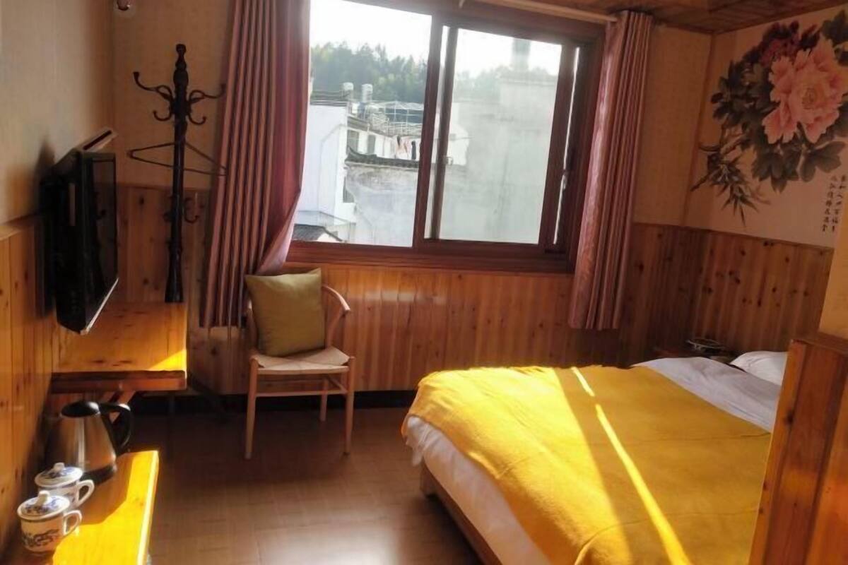 A wooden room with a bed, a chair, a table with a kettle, and a TV on the wall.