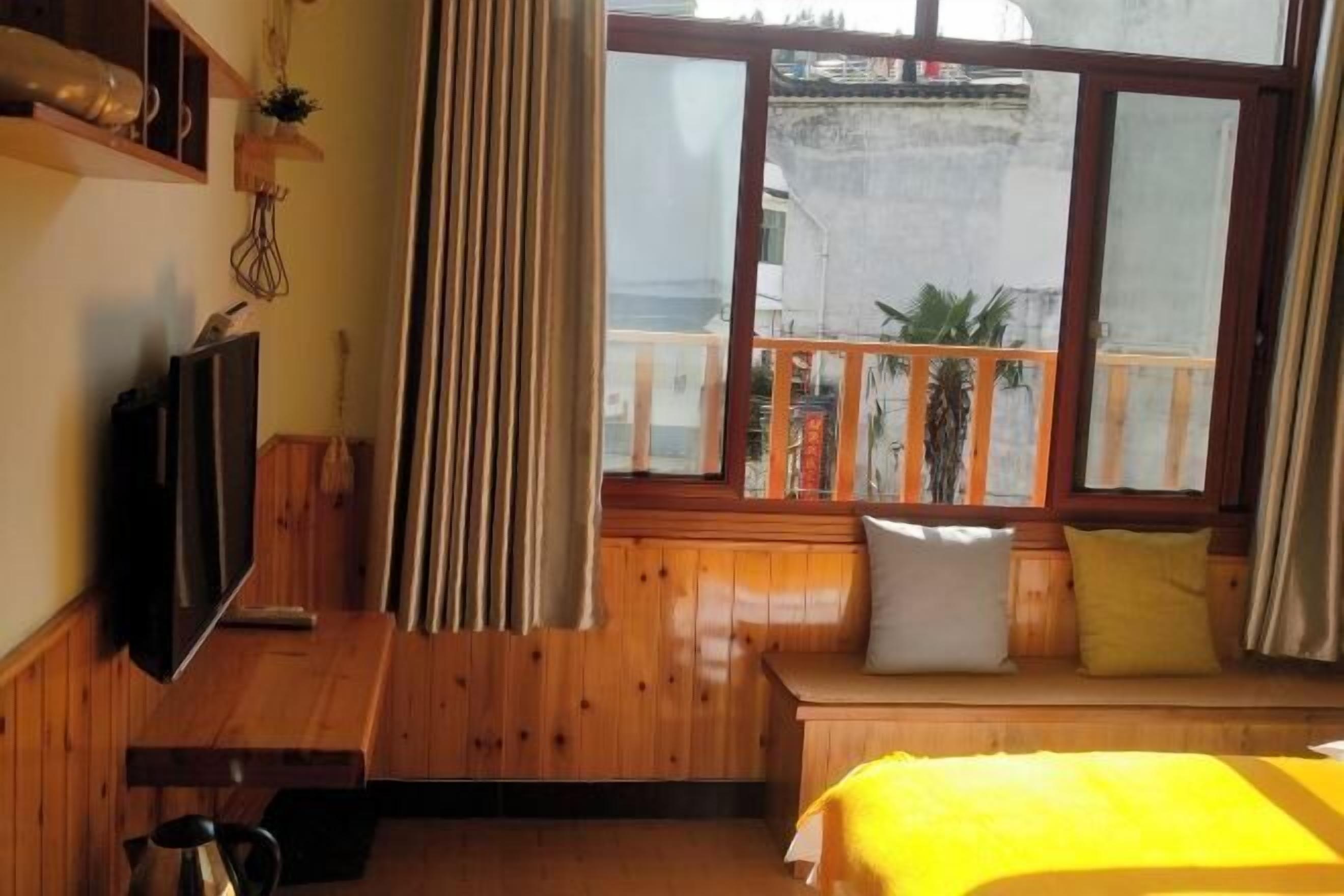 Una habitación con cama, televisor, banco de madera con cojines, ventana con vista y cortina.