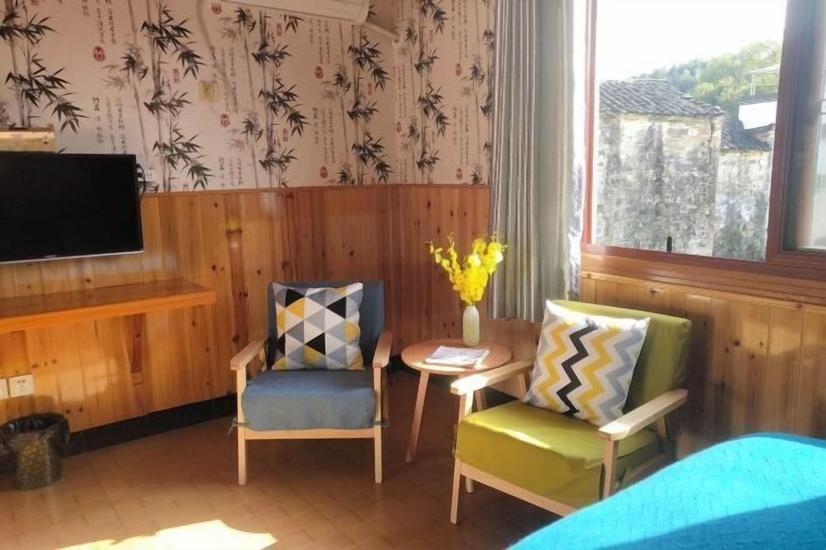 Una habitación con paneles de madera, dos sillas con cojines estampados, una mesita con un florero y una cama con cubierta azul.