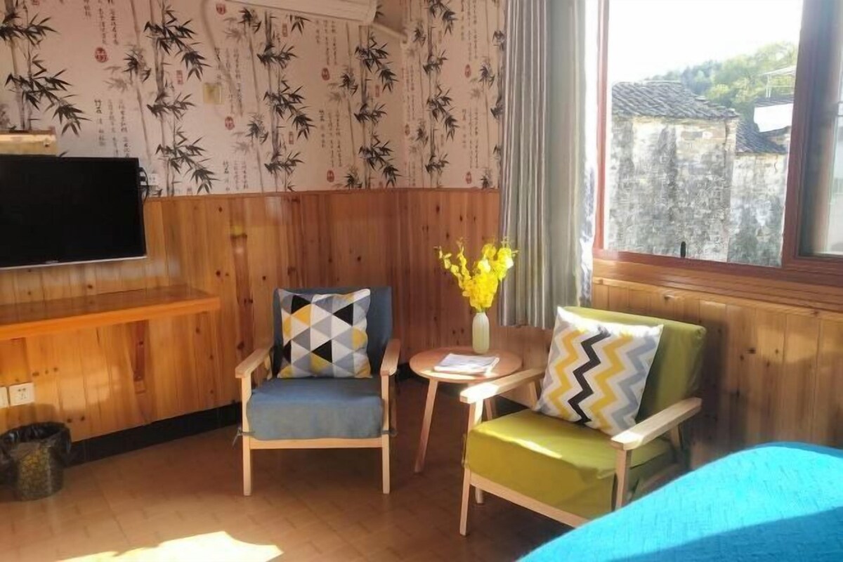 A room with wooden paneling, two chairs with patterned cushions, a small table with a vase of flowers, and a bed with a blue cover.