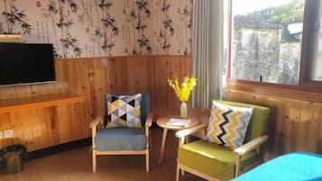 Una habitación con paneles de madera, dos sillas con cojines estampados, una mesita con un florero y una cama con cubierta azul.