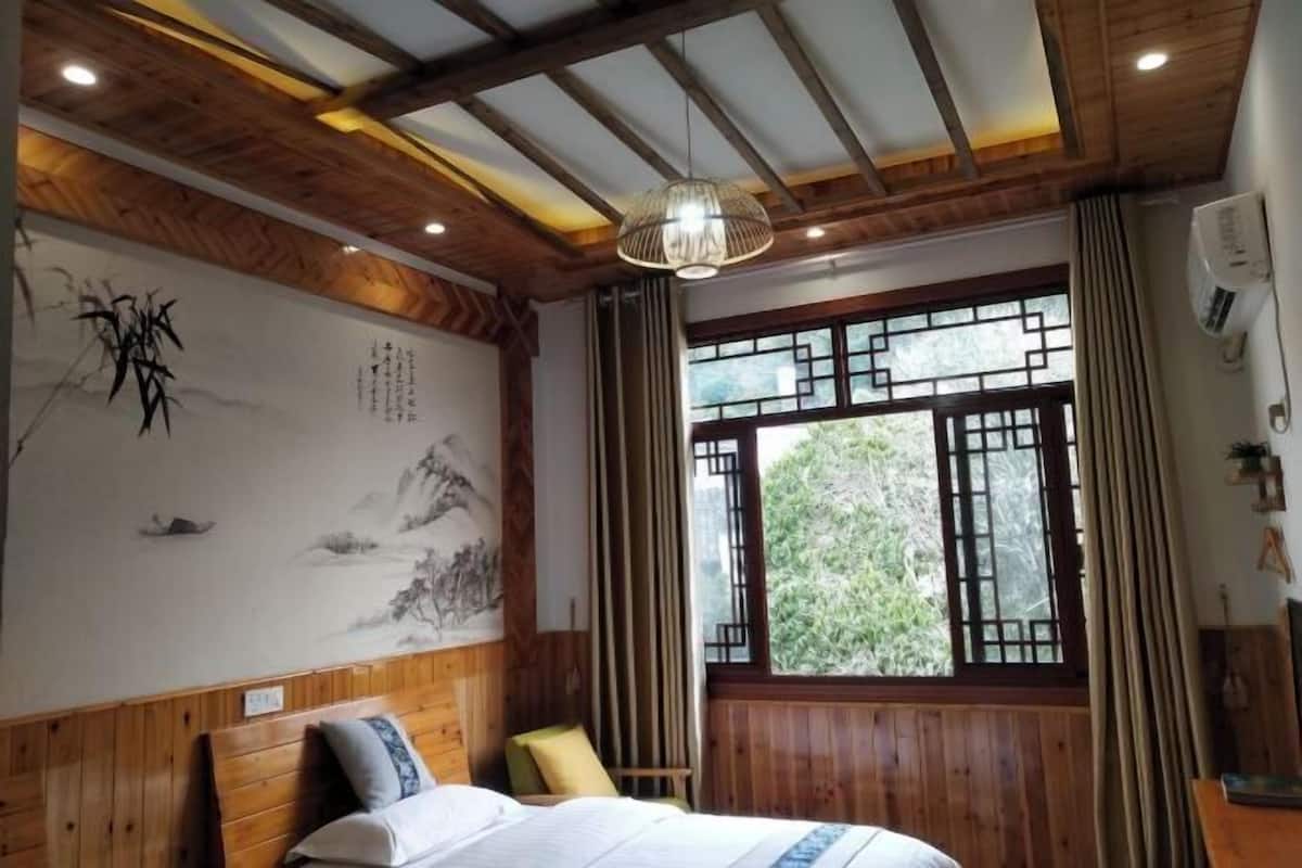 A room with wooden paneling, a bed with pillows, a hanging light fixture, a window with a view of greenery, and a wall with calligraphy.
