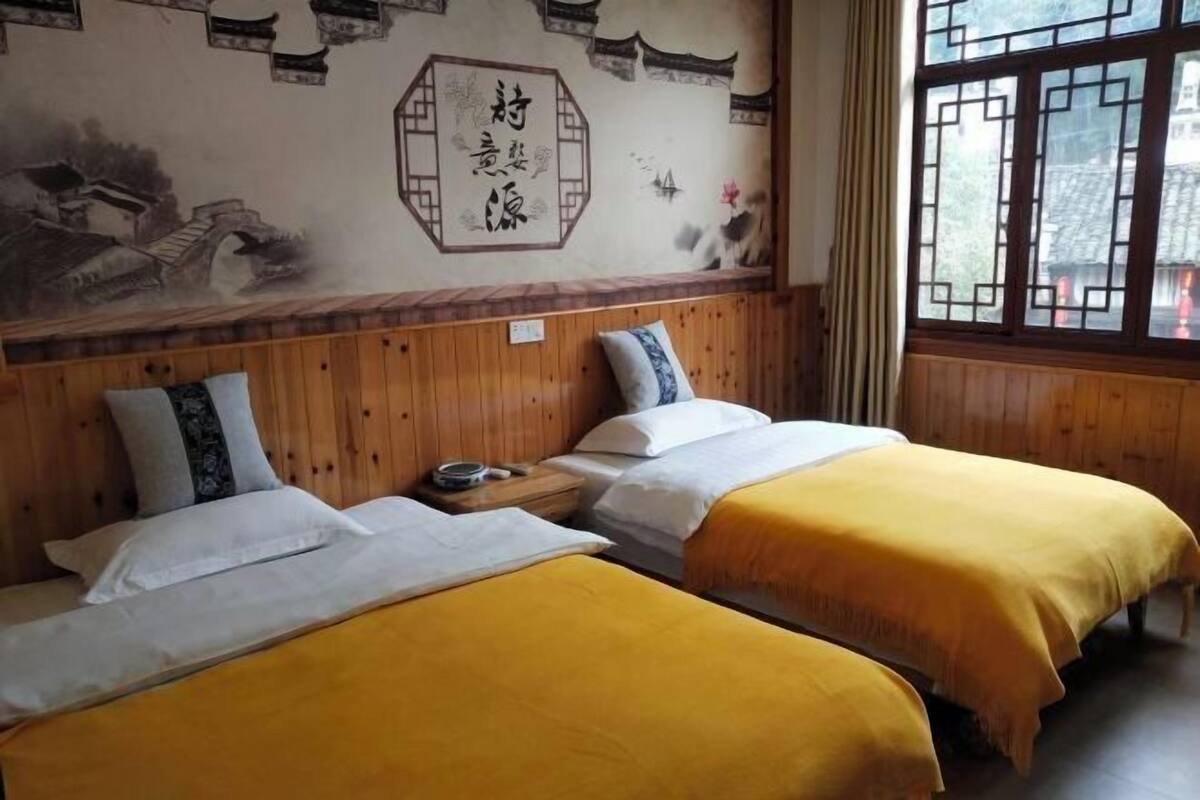 A room with two beds, wooden paneling, a traditional mural, and a window with a view.