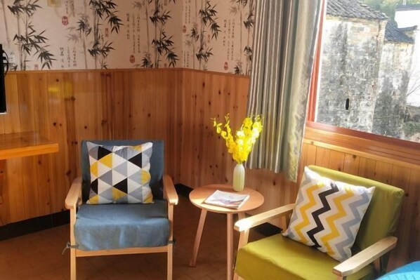 A room with wooden paneling, two armchairs with patterned cushions, a small table with a vase of flowers, and a view of a stone building through a window. - Brook Hotel (Wuyuan)
