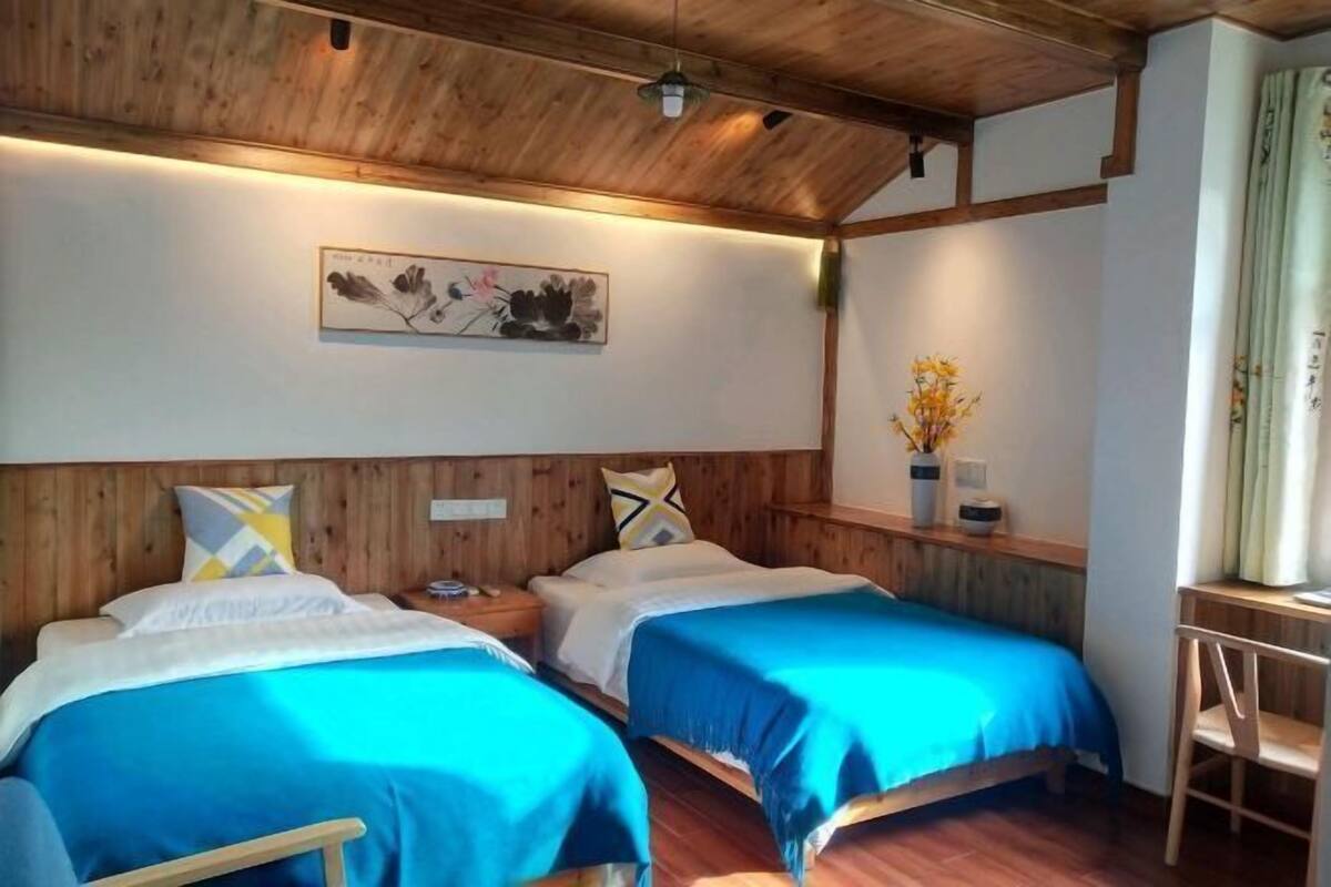 A room with two beds, wooden headboards, a wooden ceiling, a wall-mounted artwork, a bedside table with a lamp, and a window with curtains.