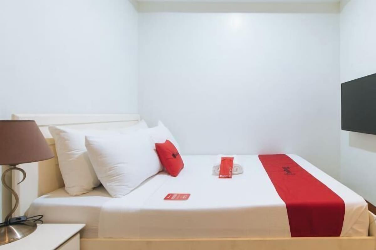 A hotel room with a bed, white walls, a red and white bedspread, a red pillow, a lamp, and a TV mounted on the wall.