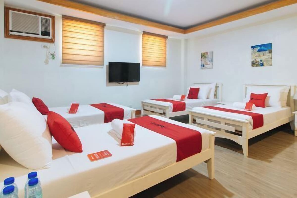 A hotel room with three single beds, each with red bedding and pillows, a television mounted on the wall, and wooden flooring.