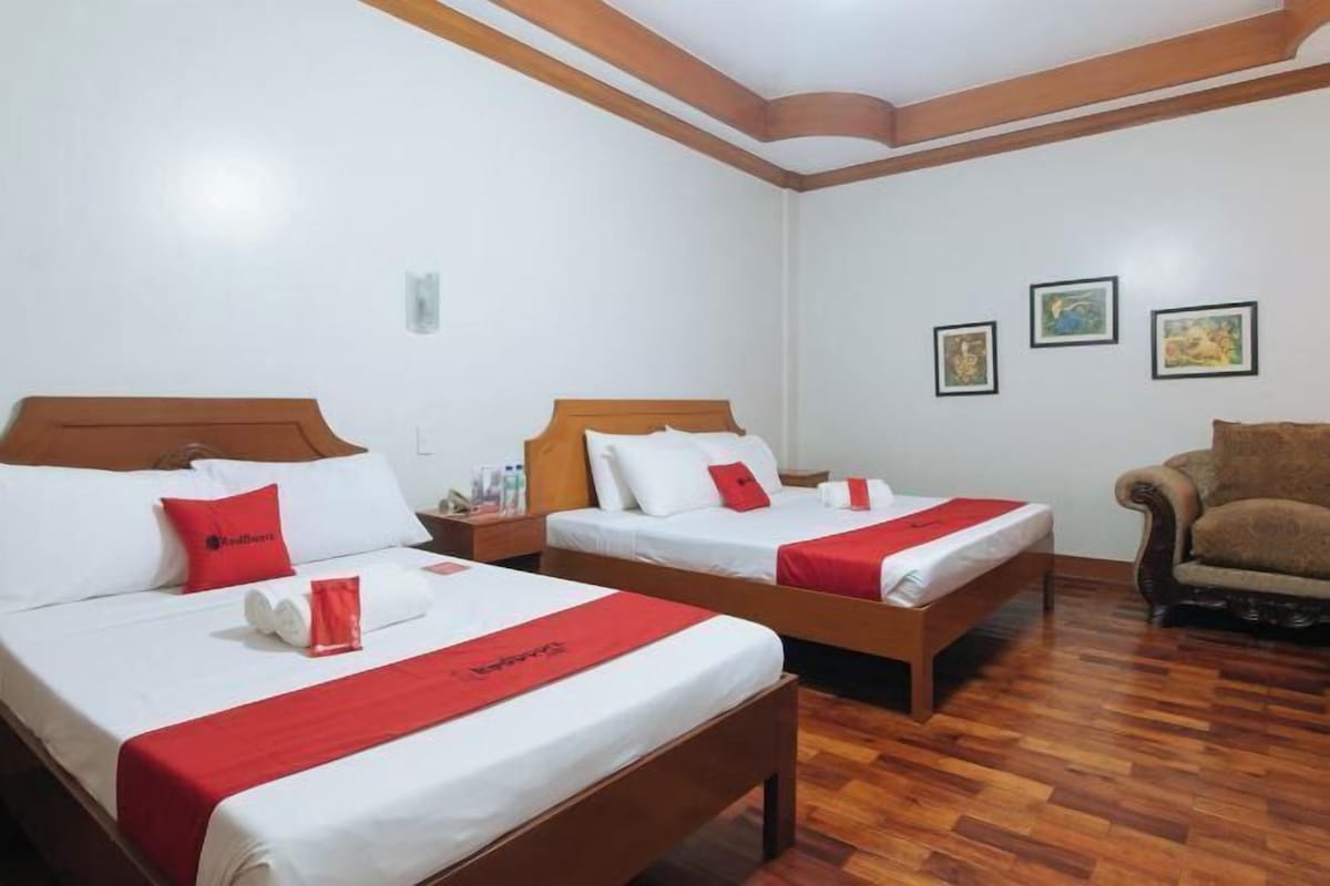 A hotel room with two beds, wooden headboards, white bedding, red accents, and a brown armchair.