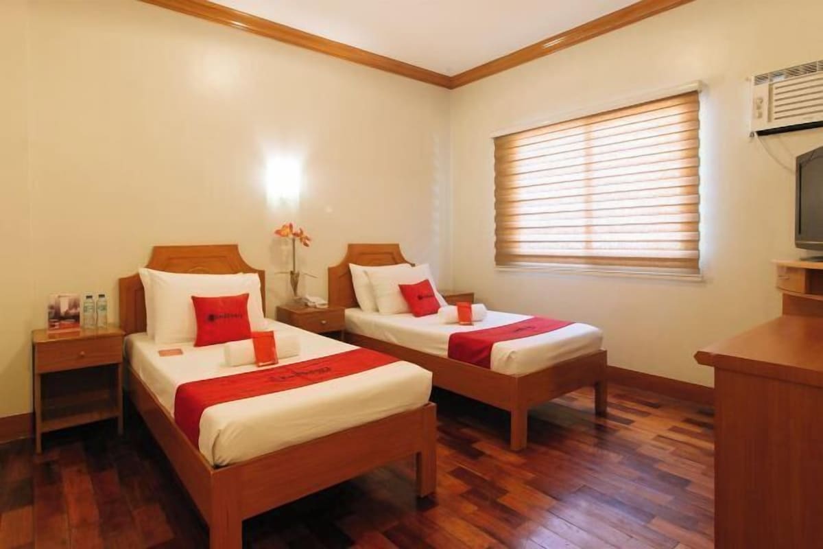 A hotel room with two single beds, wooden headboards, a nightstand with a lamp, a window with blinds, and a TV on a stand.