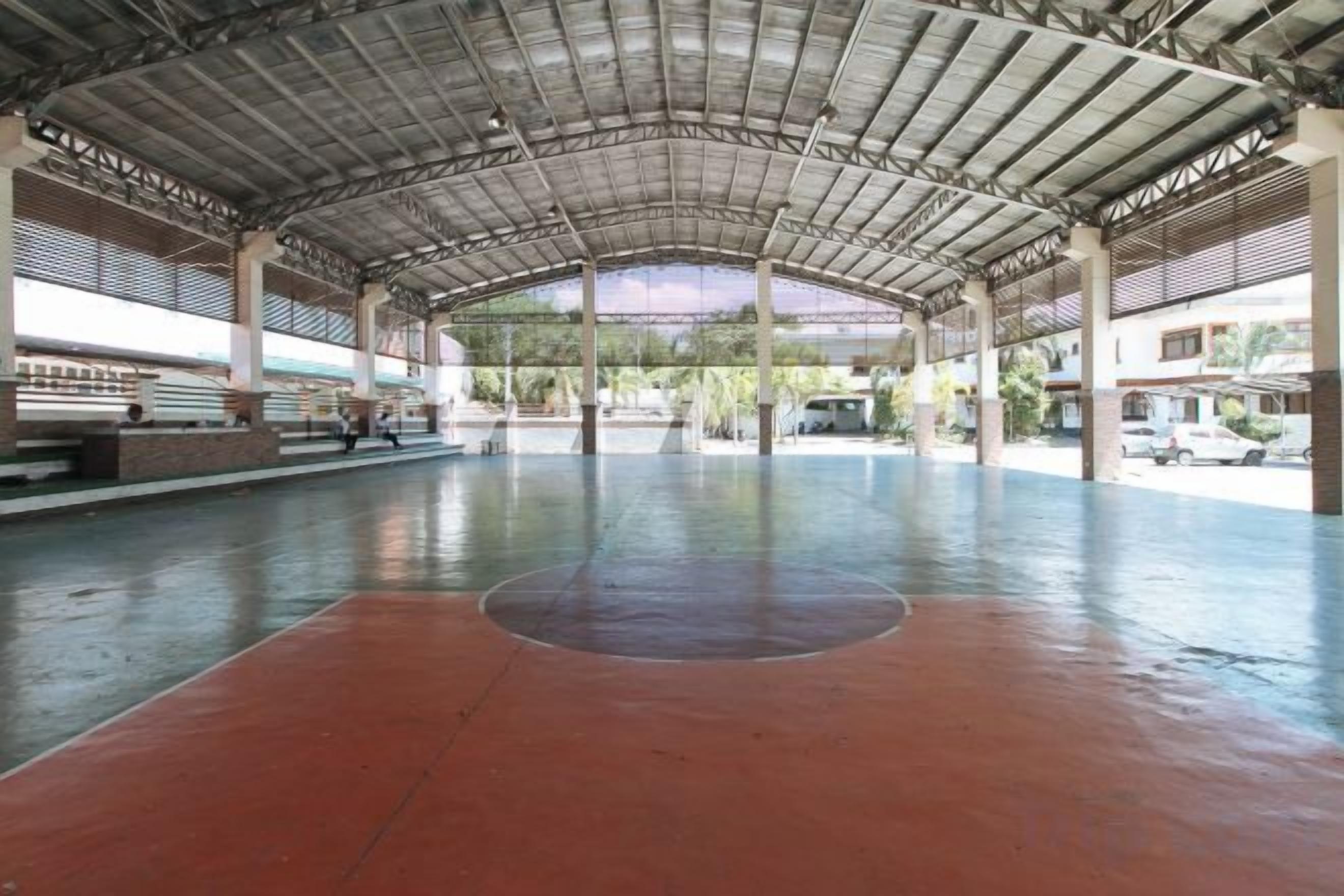 Una cancha deportiva interior con piso rojo y estructura de techo metálico.