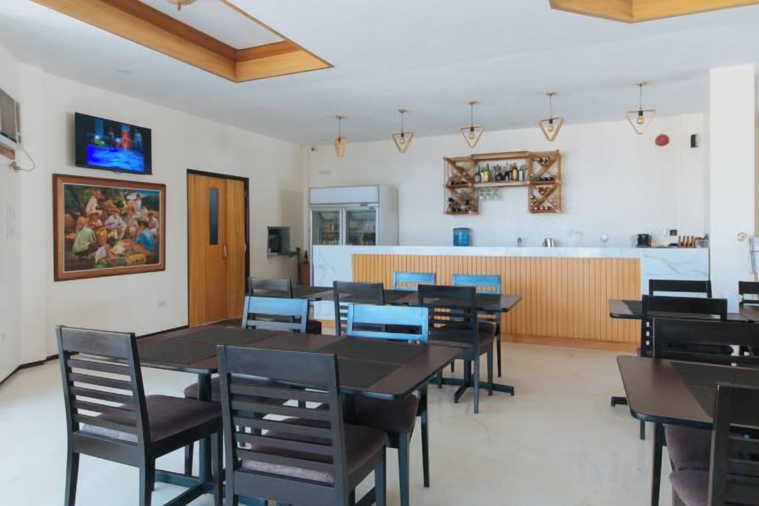 A dining area with a long table and chairs, a kitchen in the background, and a TV mounted on the wall.