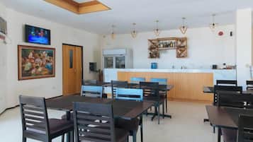 A dining area with a long table and chairs, a kitchen in the background, and a TV mounted on the wall.