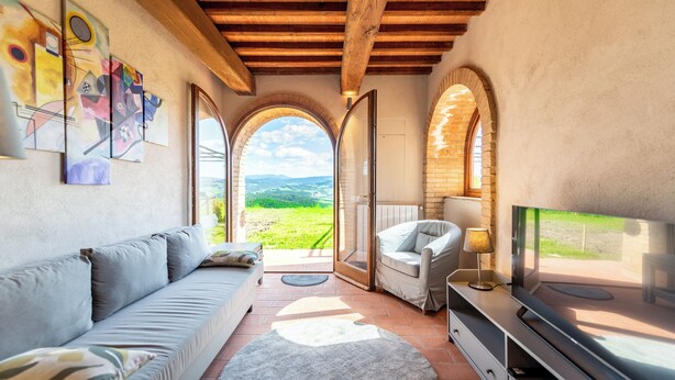 A cozy living room with a sofa, TV, and artwork on the wall. There are arched windows and a view of the countryside. - Refined Home W/ Pool, Stunning View, and Relaxing (Volterra)