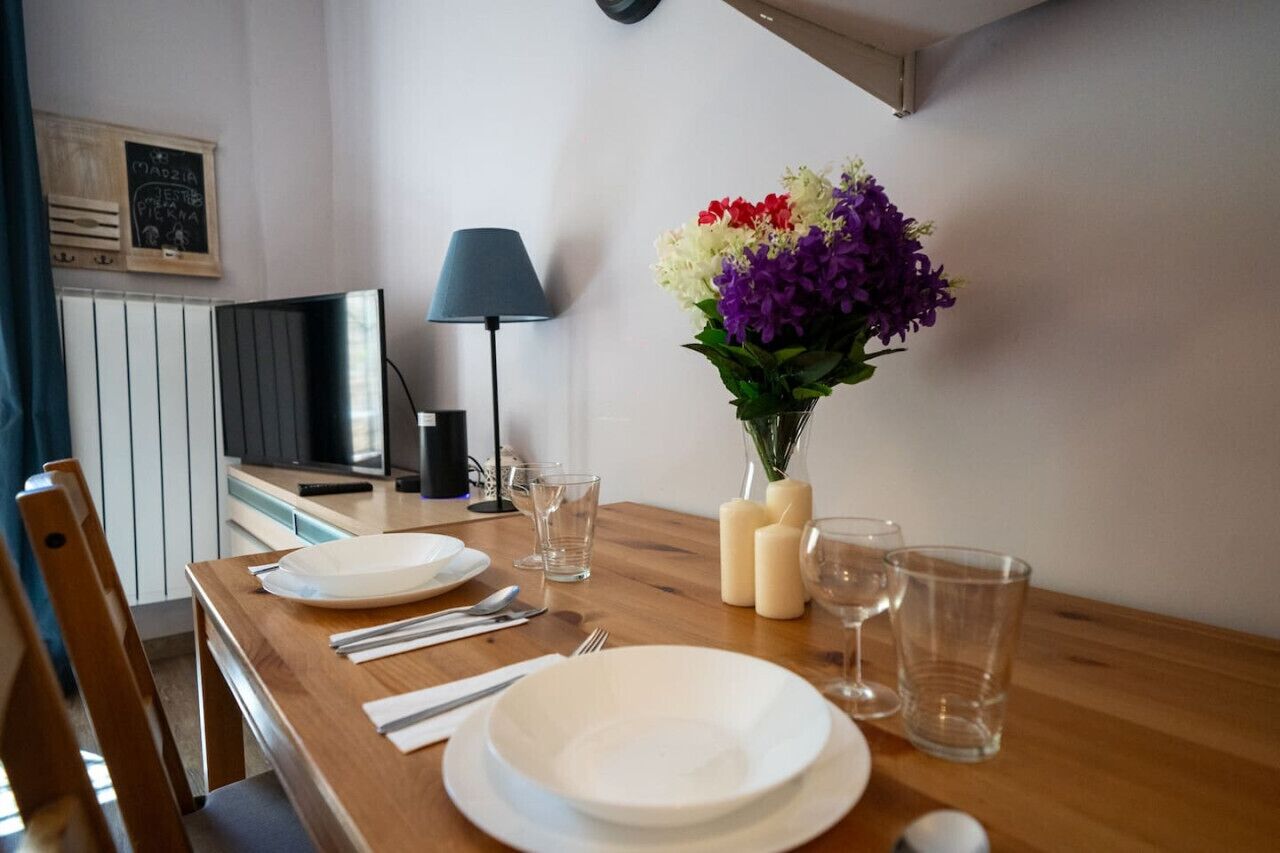A dining table set with plates, cutlery, and glasses, a vase with flowers, a lamp, and a television in a room with a window and a wall-mounted clock.