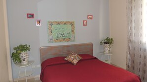 A bedroom with a red bedspread, a wooden headboard, two small side tables with potted plants, and a welcome sign on the wall.