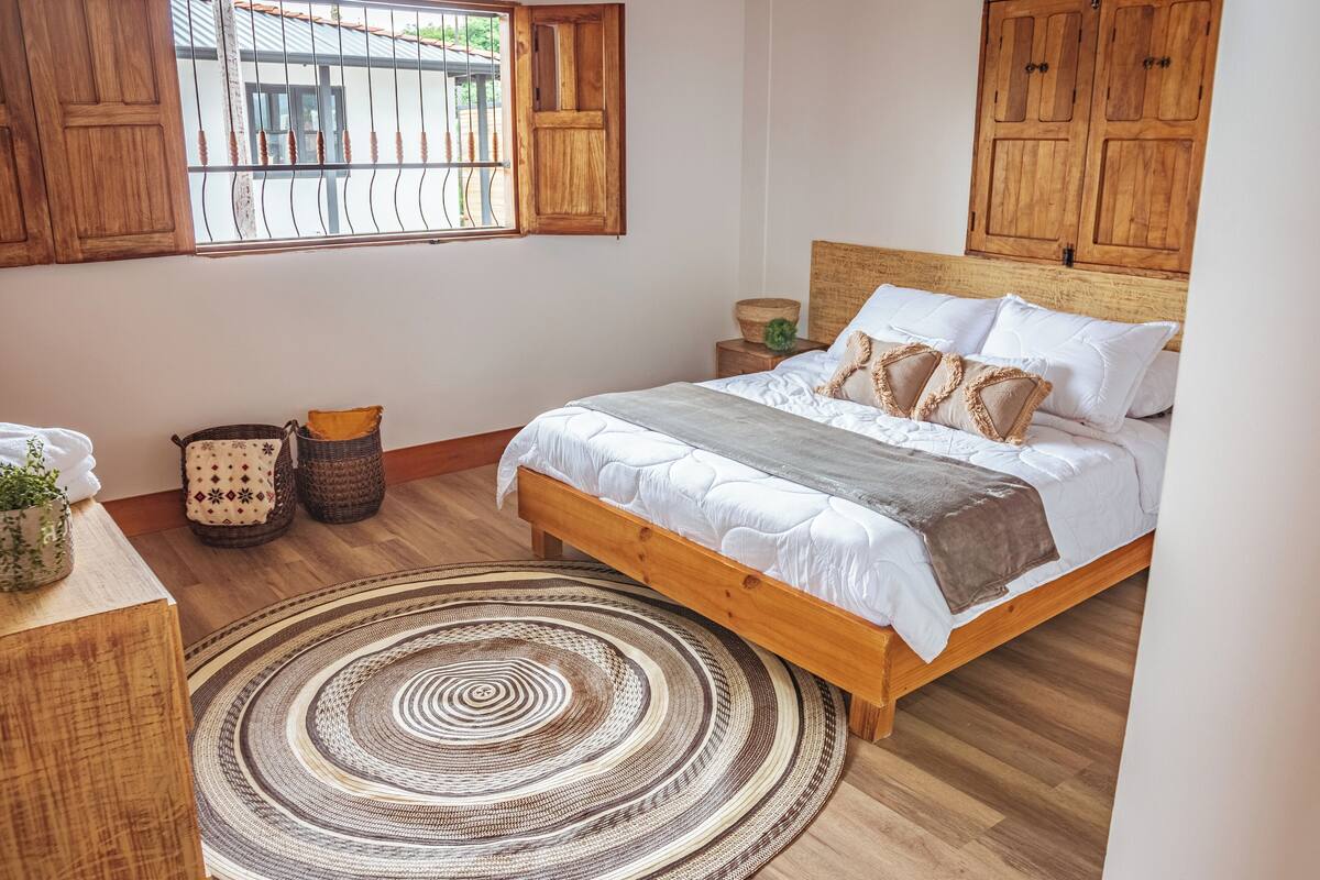A bedroom with a wooden bed, a round woven rug, and a window with wooden shutters.