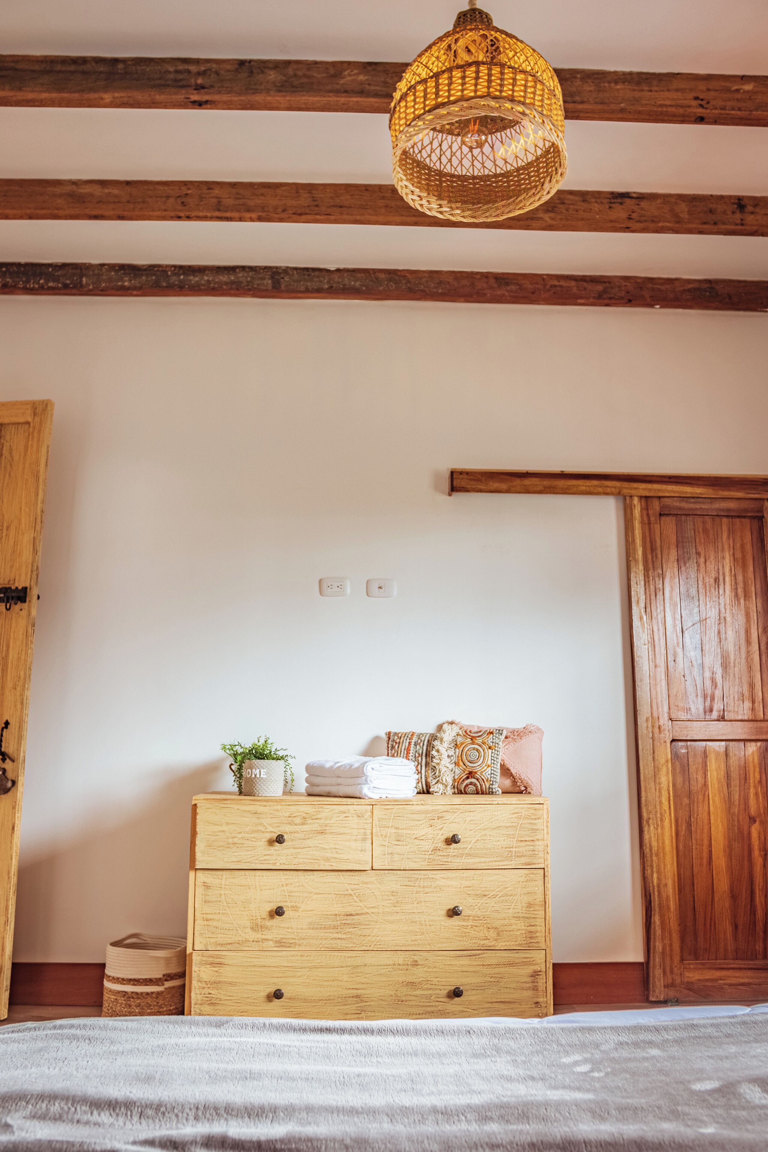 A room with exposed wooden beams, a wooden dresser with drawers, a hanging light fixture, and a wooden door.