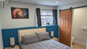 A bedroom with a bed, a nightstand, a wall-mounted lamp, a window with curtains, a door, and a painting on the wall. - The Livery (Brockton)