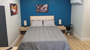 A bedroom with a bed, two bedside tables, a nightstand, a wall-mounted air conditioner, and a potted plant. - The Livery (Brockton)