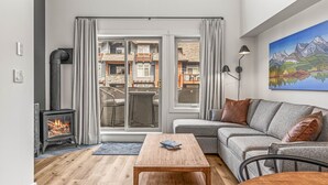 A cozy living room with a fireplace, a sofa, a coffee table, and a view of a balcony with a cityscape. - Modern Penthouse, Breathtaking Views, Hot tub/Pool (Canmore)