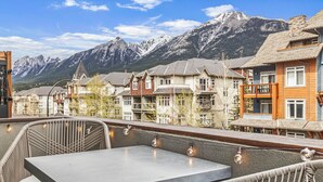 A terrace with wicker furniture and string lights, overlooking a row of traditional houses with a mountain backdrop. - Modern Penthouse, Breathtaking Views, Hot tub/Pool (Canmore)