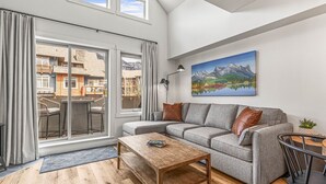 A modern living room with a grey sofa, wooden coffee table, and a painting of mountains on the wall. - Modern Penthouse, Breathtaking Views, Hot tub/Pool (Canmore)