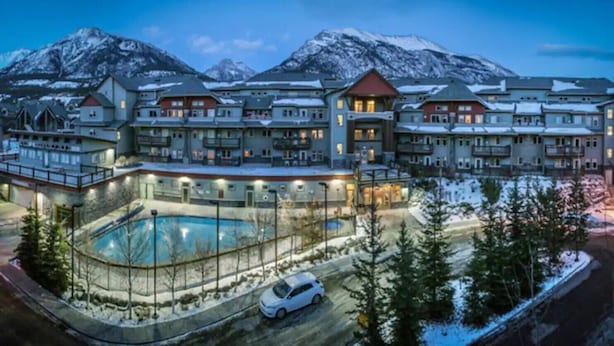 A resort with multiple buildings, a swimming pool, and snow-covered mountains in the background. - Modern Penthouse, Breathtaking Views, Hot tub/Pool (Canmore)