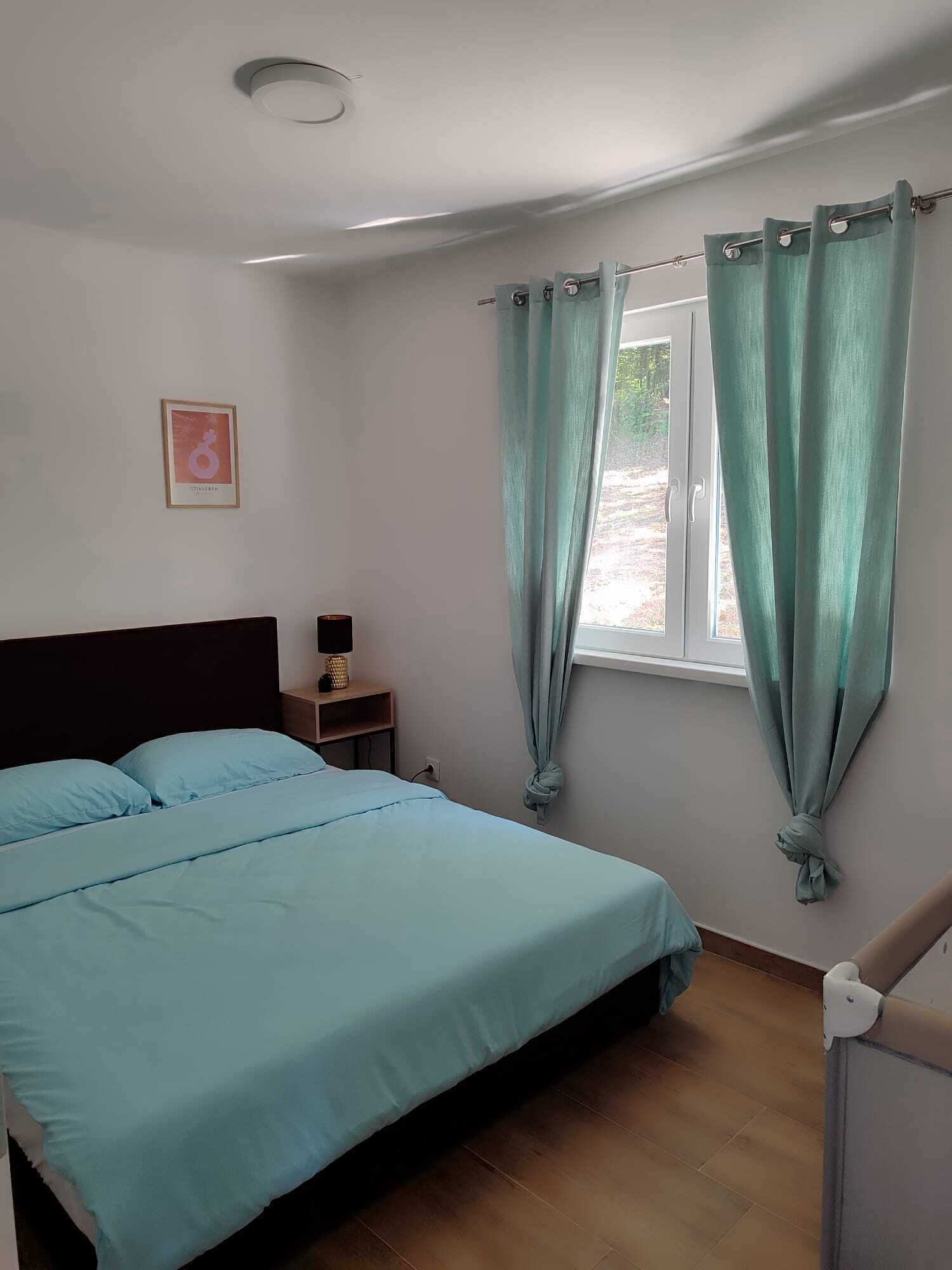 A bedroom with a bed, a nightstand, a window with curtains, and a framed picture on the wall.