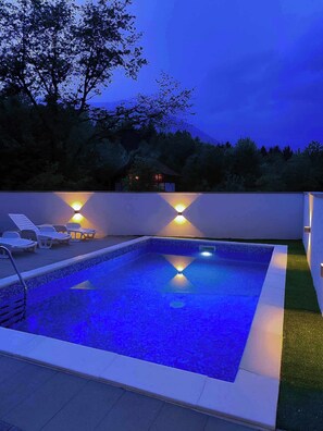 A rectangular swimming pool with a tiled edge, surrounded by a low white wall. There are lounge chairs and a small deck area with a railing. - Villa Sara Vučjak (Bihac)