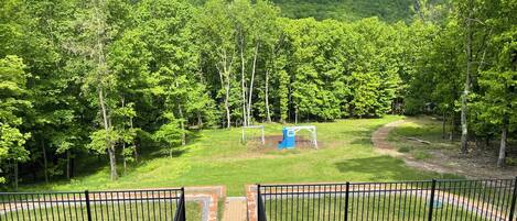 A fenced outdoor area with a playground, surrounded by dense trees and a hilly backdrop.