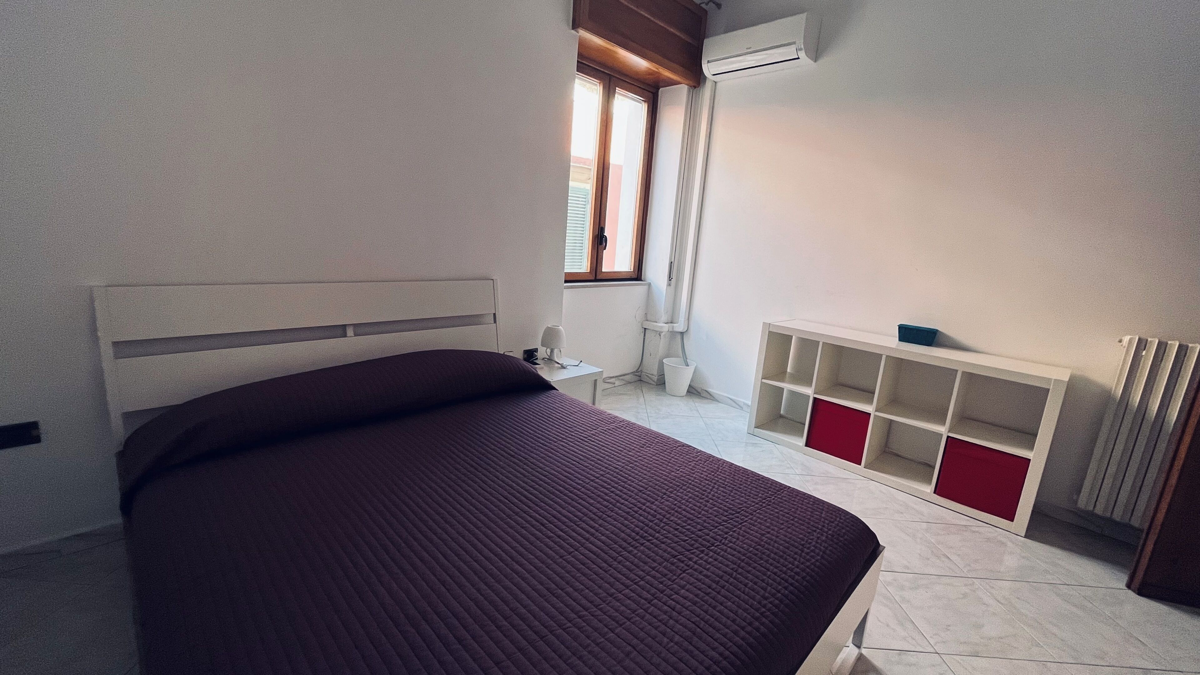 A bedroom with a bed, a radiator, a window, and a white shelf unit.