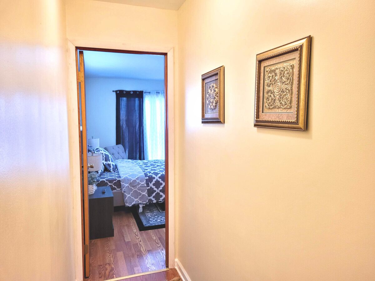 Corredor que dá para um quarto com cama, mesa de cabeceira e cortinas.