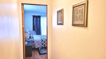 Corredor que dá para um quarto com cama, mesa de cabeceira e cortinas.