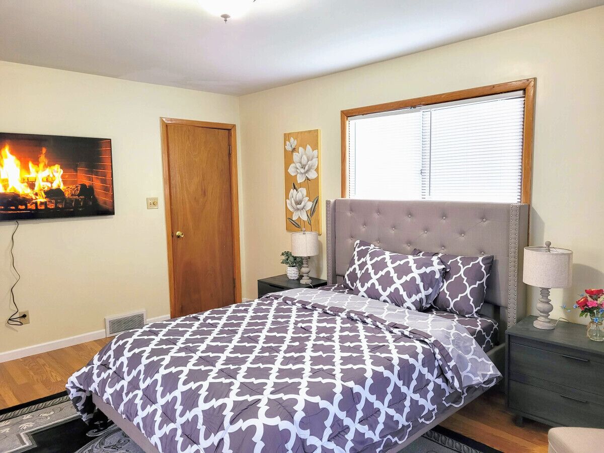 A bedroom with a bed, a nightstand, a wall-mounted TV, a door, and a window with blinds.