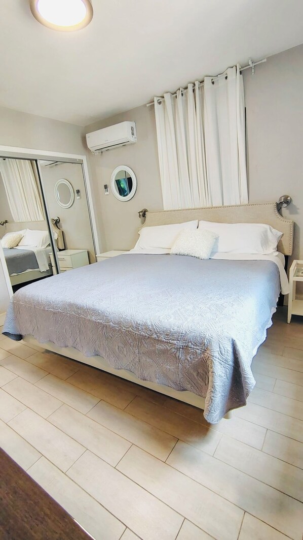 Un dormitorio con cama, mesita de noche, espejo, equipo de aire acondicionado y ventana con cortinas.