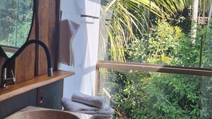 A bathroom with a wooden vanity, a round mirror, and a sink. There is a window with a view of greenery outside. - Pousada Portaldo Céu  (Domingos Martins)