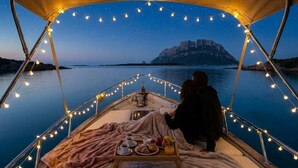 A boat with a covered deck, string lights, a table with food and drinks, and a view of a rocky coastline at dusk. - Wild Escape Island Bay Experience (Sardegna)