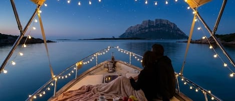 A boat with a covered deck, string lights, a table with food and drinks, and a view of a rocky coastline at dusk.