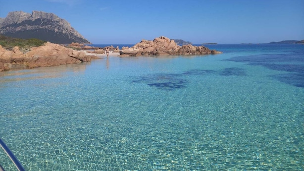 A clear coastal water body with rocky formations and a distant mountain. - Wild Escape Island Bay Experience (Sardegna)