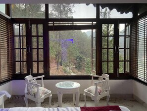 A room with a large bay window, two white chairs, a small round table, and a bed with a red blanket. - Luxsugero Penthouse Kaichidham (Nainital)